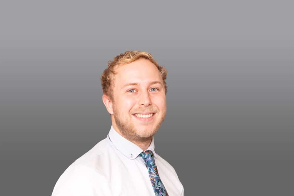 A man with short blond hair and a beard smiles at the camera in a style reminiscent of a Birmingham commercial photographer. He is wearing a white shirt and a patterned tie, with a plain grey background behind him.