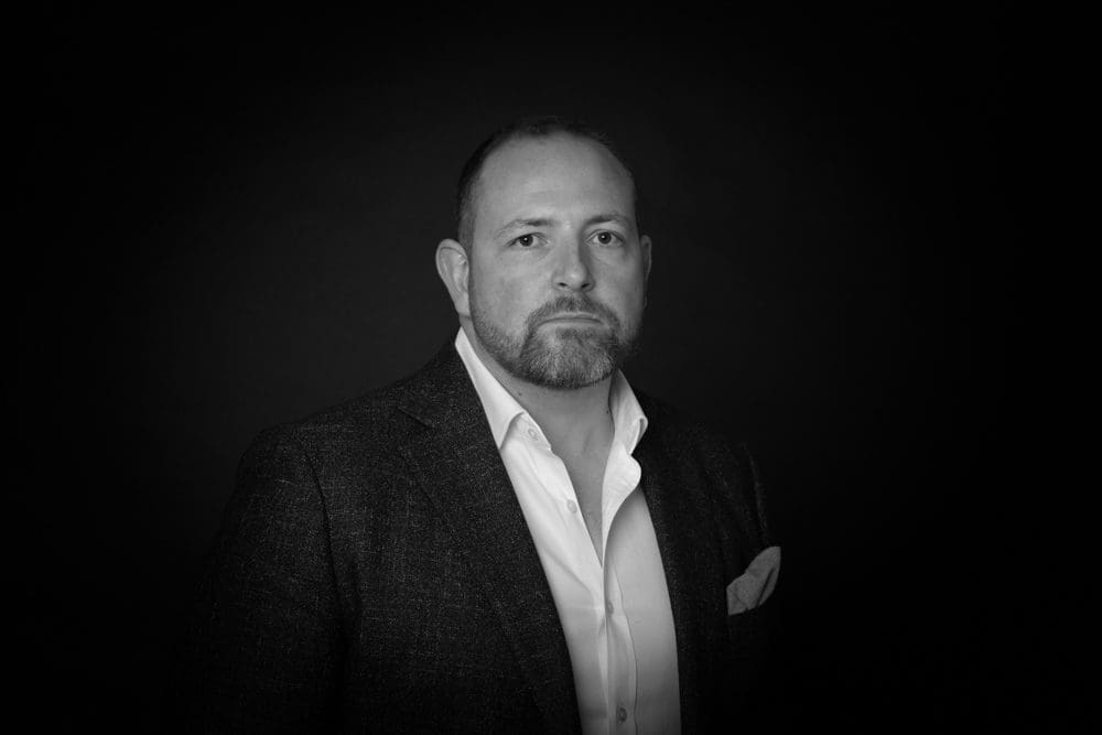 A man with a beard and short hair, wearing a dark suit jacket and a white shirt, poses against a dark background in a black-and-white portrait by a Birmingham commercial photographer.