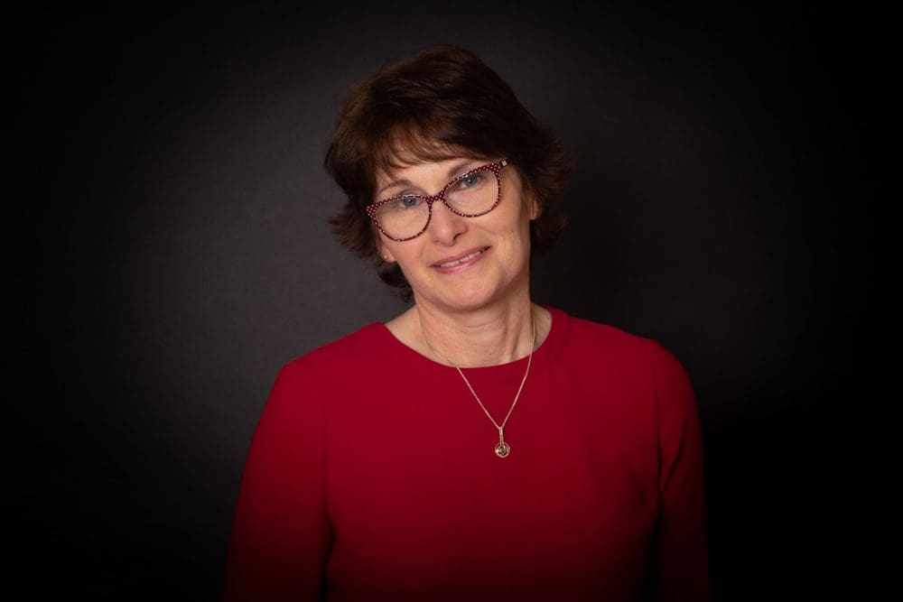 A woman with short brown hair and glasses, wearing a red top and a necklace, stands in front of a dark background, smiling gently at the camera—captured with the professional touch of a Birmingham commercial photographer.