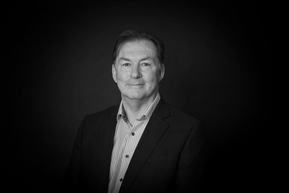 Black and white portrait of a middle-aged man in a suit jacket and striped shirt, smiling slightly, with a dark, plain background—captured with the expertise of a Birmingham commercial photographer.