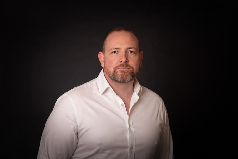 A man with short hair and a beard wears a white button-up shirt and stands against a plain black background, facing the camera with a neutral expression in the style of a Birmingham commercial photographer.
