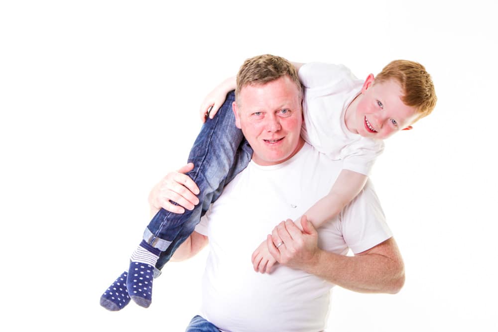 A man in a white shirt carries a smiling young boy over his shoulders. The boy wears a white shirt, blue jeans, and spotty socks. Both are against a plain white background.