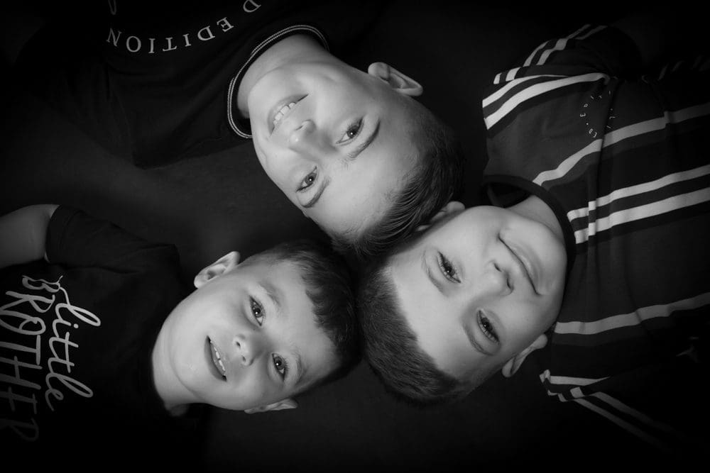 Three young boys lying on their backs in a circle, heads touching, smiling up at the camera. The photo is in black and white.
