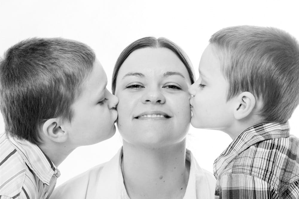 A woman smiles as two young boys, one on each side, kiss her cheeks. The image is in black and white.