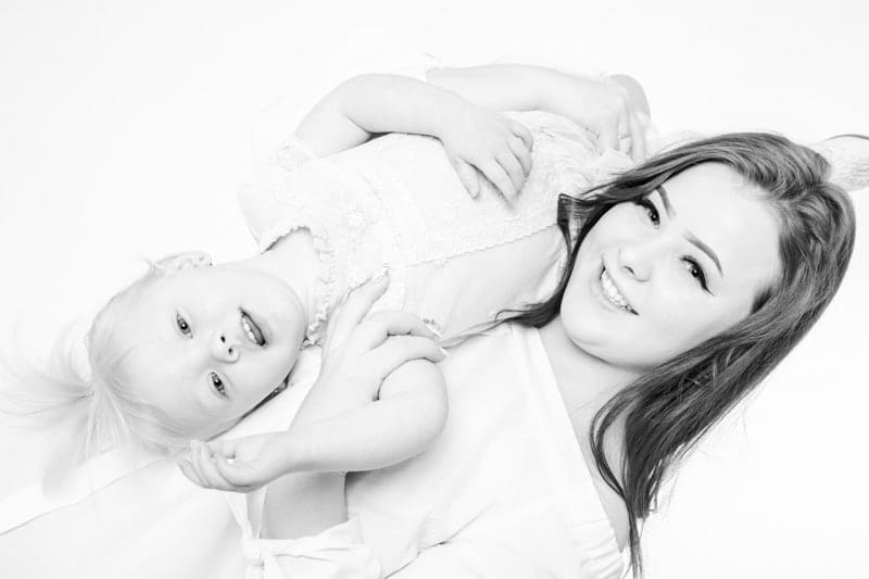 A woman lies on her back, smiling at the camera, whilst holding a young child who is lying on top of her and looking up. Both are dressed in light clothing against a white background.
