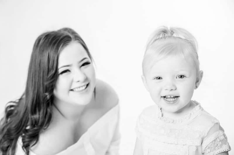 A woman with long hair and a smiling young girl with fair hair in a ponytail pose together against a plain white background.
