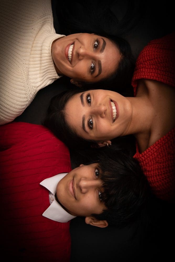 Three people lie on a dark background with their heads touching, forming a triangle. Two wear red jumpers, one in a white polo neck. All are smiling and looking up at the camera—capturing the warmth and connection that a Birmingham family photographer is known for.