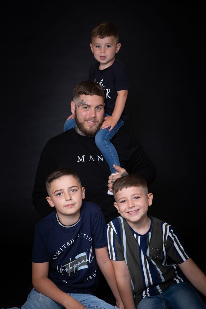 Four people pose against a black background: an adult man in a black jumper with three young boys, one sitting on his shoulders and two sitting in front, all smiling.
