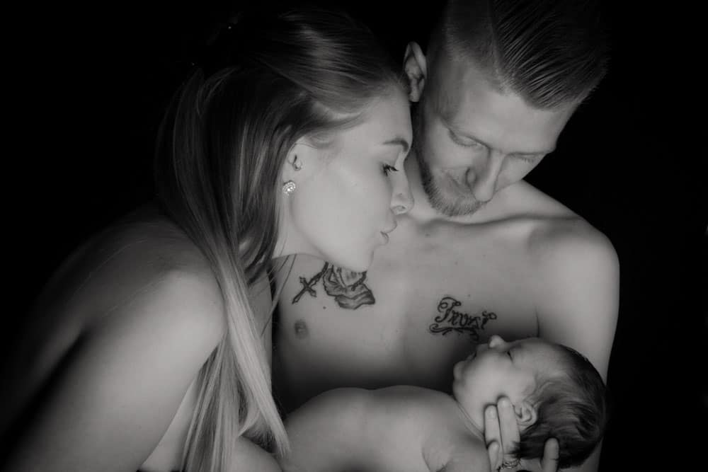 A black and white photo of a woman and man lovingly holding a newborn baby, with both adults looking down at the infant. The father's chest tattoos are visible.