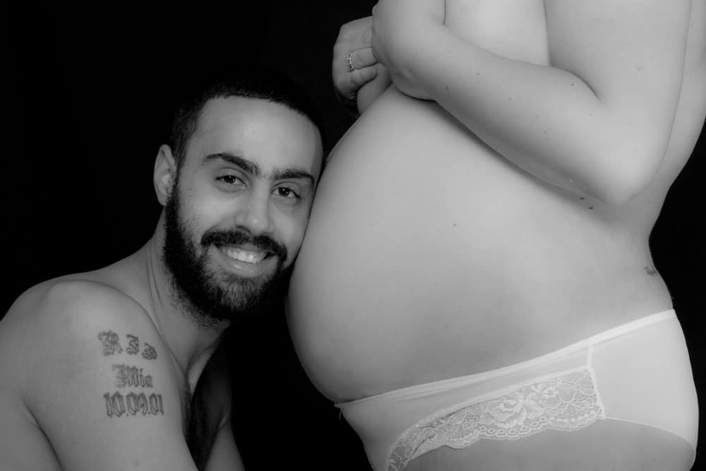 A smiling man with a beard rests his head against a pregnant woman's bare belly, both posed against a black background.