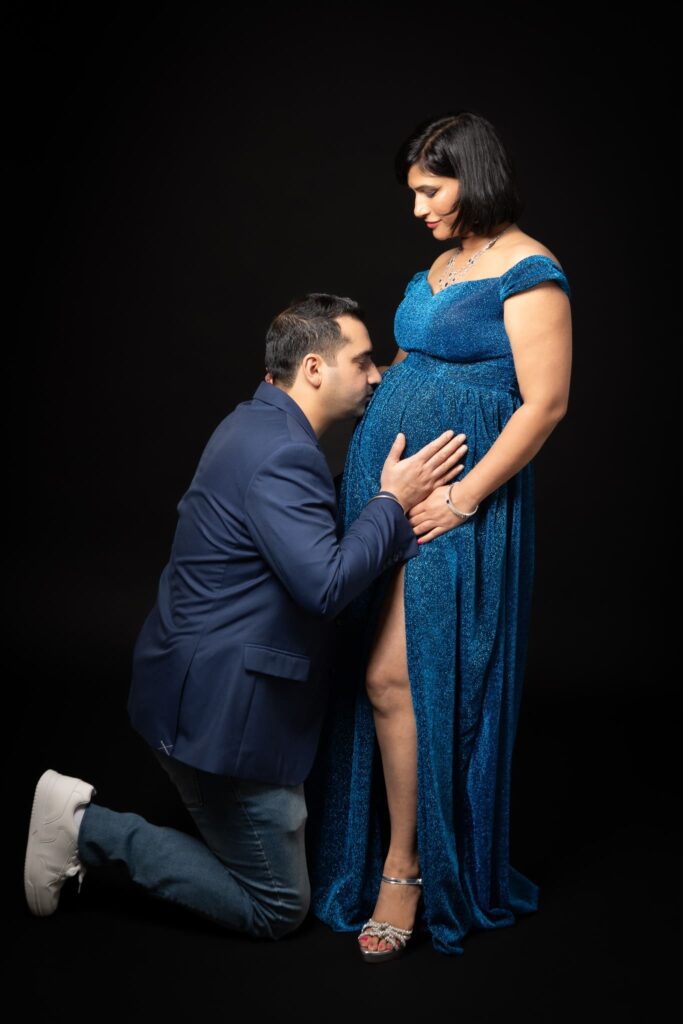 A man kneels, gently holding and kissing a pregnant woman's belly. The woman stands wearing a sparkly blue gown, smiling down at him against a black background.