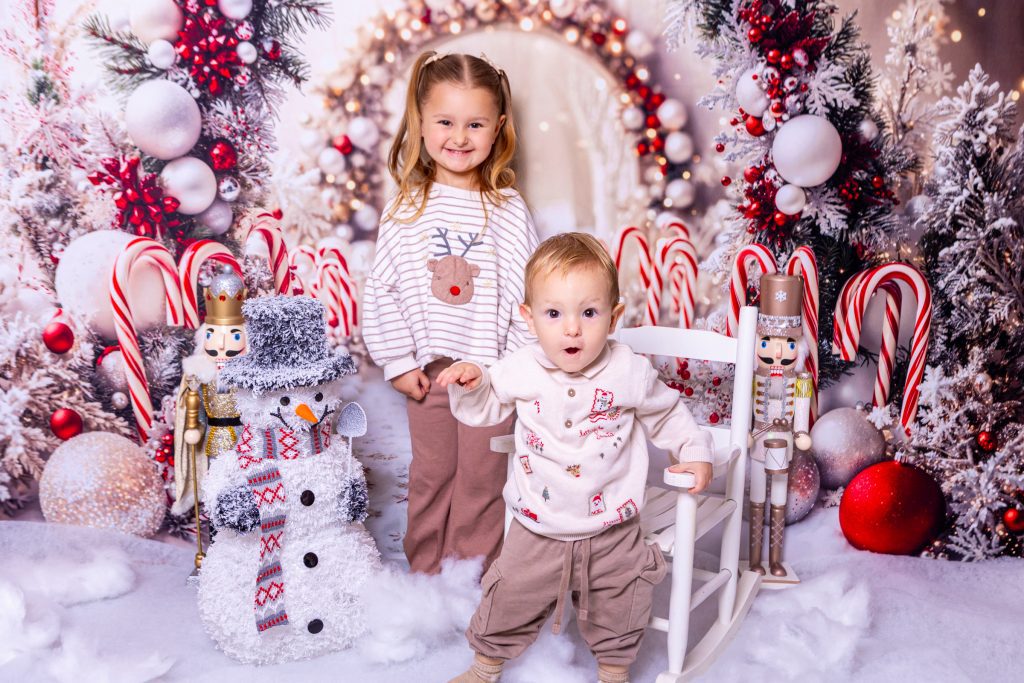 Two young children pose in a festive, snowy Christmas scene with candy canes, nutcrackers, baubles, and a smiling snowman. Captured by a talented Birmingham photographer, the older child stands behind the younger, who holds a small white rocking chair.