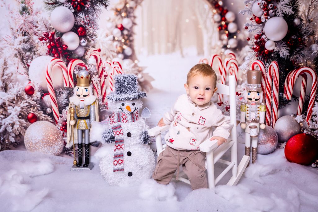 A baby in a white chair smiles, surrounded by nutcracker figures, a snowman, candy canes, red baubles, and snowy holiday decorations in a festive winter scene captured by a Birmingham photographer.