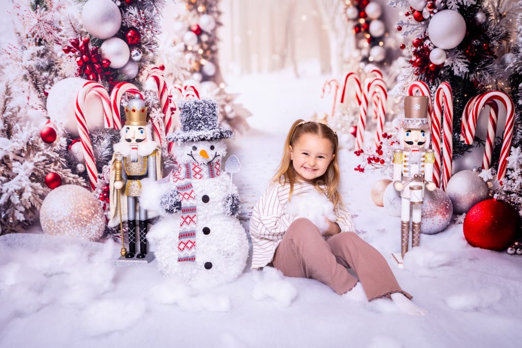 Young girl smiles sitting on a snowy floor, surrounded by two nutcracker figures, a snowman, red baubles, candy canes, and festive winter decorations. Captured beautifully by a Birmingham photographer, the scene comes alive against a backdrop filled with white trees and glistening snow.
