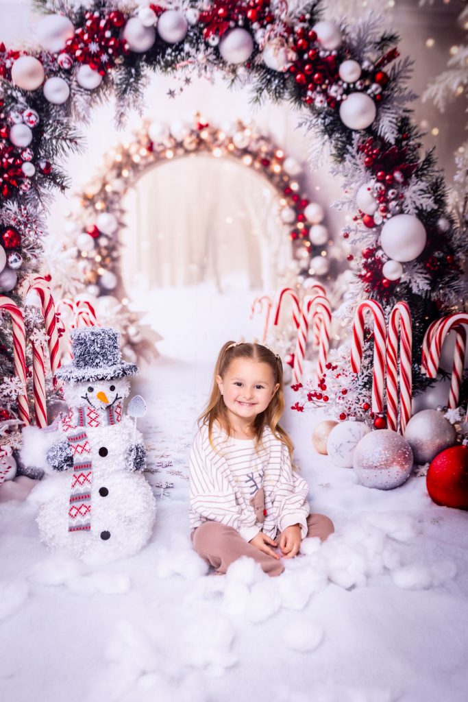 A young girl sits smiling on artificial snow in front of a festive arch with red and white baubles, candy canes, and a snowman, creating a cheerful holiday scene perfectly captured by a Birmingham photographer.