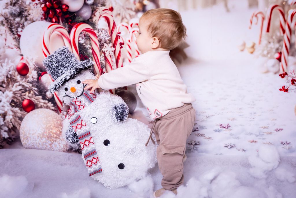 A toddler stands in the snow, touching a snowman with a scarf and hat, beside a decorated Christmas tree with candy canes and baubles, beautifully captured by a Birmingham photographer.