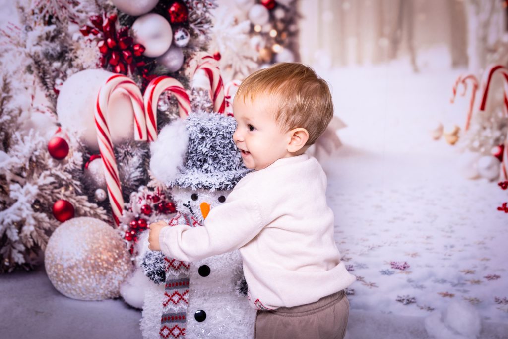 A small child in a white jumper hugs a snowman decoration in front of a festive, snowy background with Christmas trees, candy canes, and baubles—captured beautifully by a Birmingham photographer.