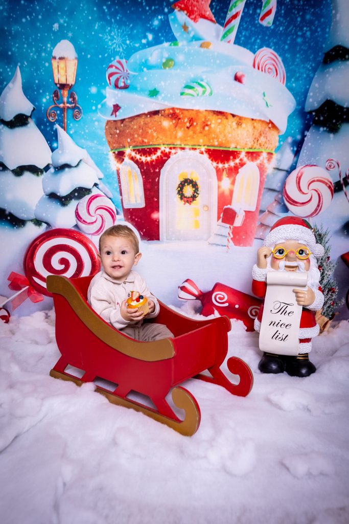 A baby sits in a red sleigh holding a cupcake, surrounded by artificial snow and Christmas decorations, with a festive gingerbread house and Father Christmas holding The Nice List in the background—captured perfectly by a talented Birmingham photographer.