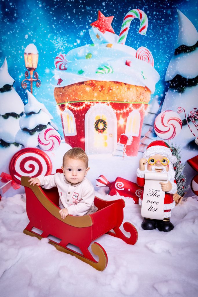 A baby sits in a red sleigh on a snowy set with sweets and gingerbread house decorations, beautifully captured by a Birmingham photographer. A Father Christmas figure holds a sign reading The nice list. The scene is colourful and festive.
