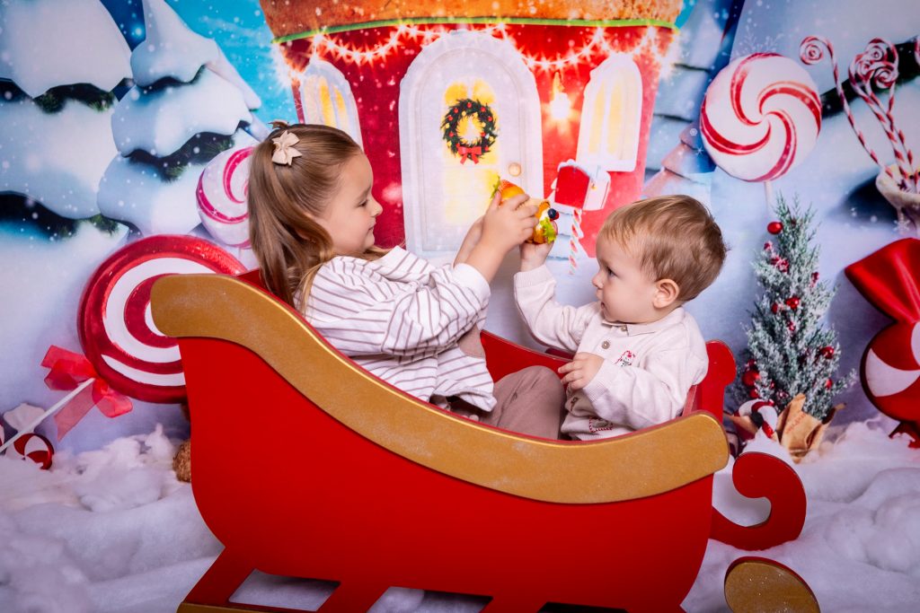 Two young children sit in a red sleigh, smiling and playing with toys. Captured by a talented Birmingham photographer, the background is decorated with candy canes, snow, and a festive gingerbread house, creating a cheerful holiday scene.