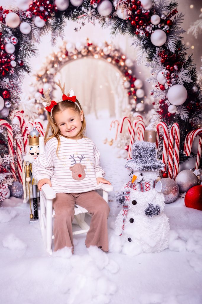 A young girl with a reindeer headband sits on a white chair in a festive, winter-themed setting with candy canes, baubles, a snowman, and Christmas decorations surrounding her—captured beautifully by a talented Birmingham photographer.