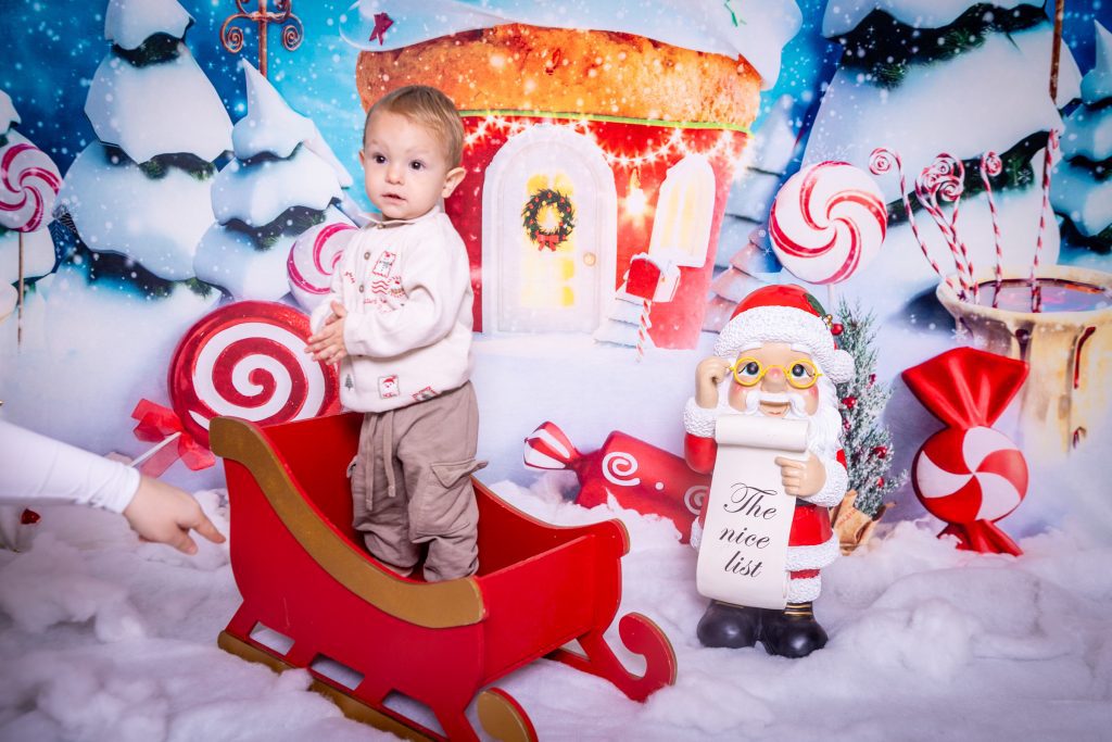 A toddler stands in a red sleigh surrounded by artificial snow, sweet decorations, and a Father Christmas figure holding a “The Nice List” scroll during a mini Christmas photoshoot in Birmingham. A hand points from the left, and a gingerbread house backdrop completes the festive scene.