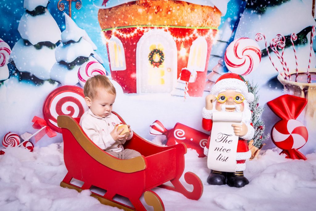 A baby sits in a small red sleigh holding a bauble, next to a Father Christmas figure holding The Nice List scroll, with a sweet-themed winter backdrop featuring a decorated house and snow—the perfect scene for a mini Christmas photoshoot in Birmingham.