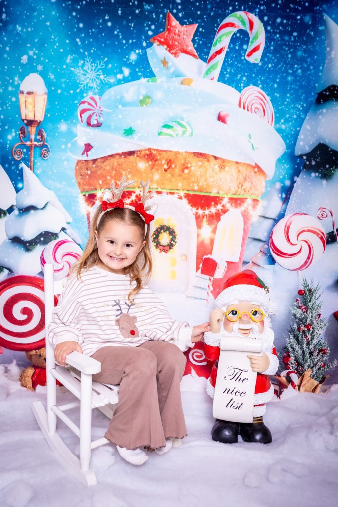 A smiling child in reindeer antlers sits on a white rocking chair next to a Santa figurine holding a The Nice List sign, with a festive sweet-themed holiday backdrop behind them—a perfect scene for a mini Christmas photoshoot in Birmingham.