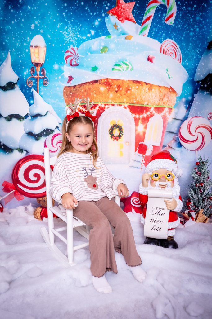 A young girl sits on a white chair in a festive, snowy setting with sweet decorations and a Father Christmas figure holding a The Nice List scroll, in front of a whimsical gingerbread house backdrop—perfect for capturing memories at a mini Christmas photoshoot Birmingham.