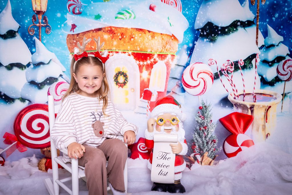 A young girl in reindeer antlers smiles whilst sitting on a white chair next to a Father Christmas figurine holding a The nice list scroll, surrounded by festive sweet decorations and snowy holiday scenery—perfect for a magical mini Christmas photoshoot in Birmingham.