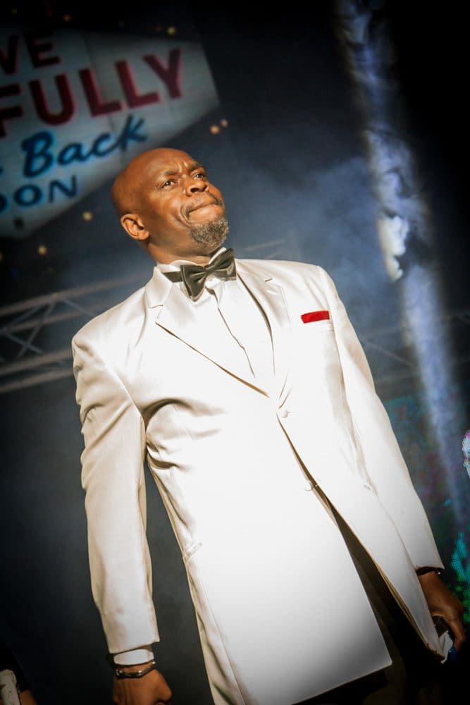 A man in a white tuxedo with a black bow tie stands on stage under dramatic lighting, with a sign and misty background behind him, perfectly captured by a skilled Birmingham event photographer.