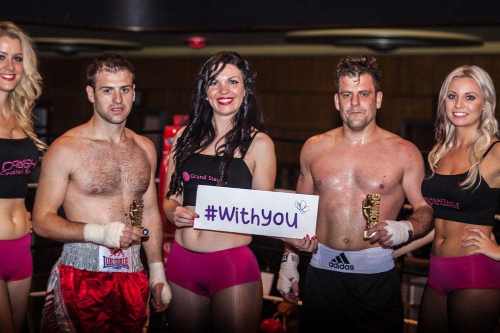 Two male boxers and three women pose together indoors, captured expertly by a Birmingham event photographer. The central woman holds a sign reading #WithYou. The boxers are bare-chested and wearing gloves, while the women wear matching black tops and pink shorts.