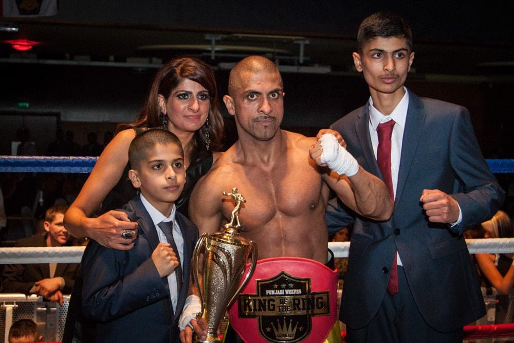 A boxer with a championship belt and trophy poses in a boxing ring with three family members—two boys in suits and a woman—all smiling for the photo after a bout, beautifully captured by a Birmingham event photographer.