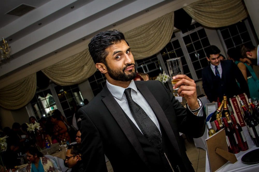 A man in a black suit holds up a drink and smiles at a formal event, with people and bottles in the background, perfectly captured by a Birmingham event photographer.