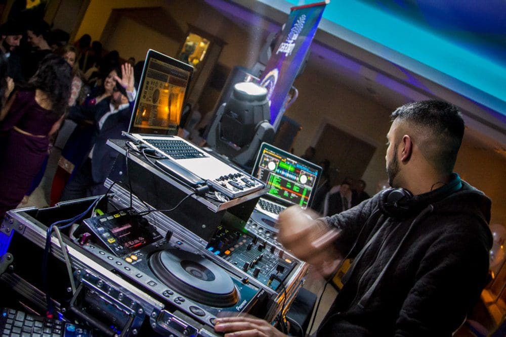 A DJ is performing at a lively event, surrounded by audio equipment, turntables, laptops, and colourful lights, whilst people dance and mingle in the background—perfect moments for a Birmingham event photographer to capture the energy and excitement of the night.