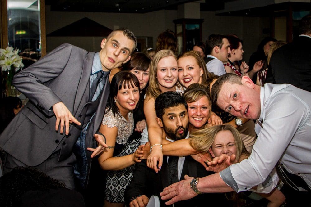 A group of people dressed formally, smiling and posing closely together at a lively indoor event, with some making playful gestures towards the camera—captured brilliantly by a Birmingham event photographer.