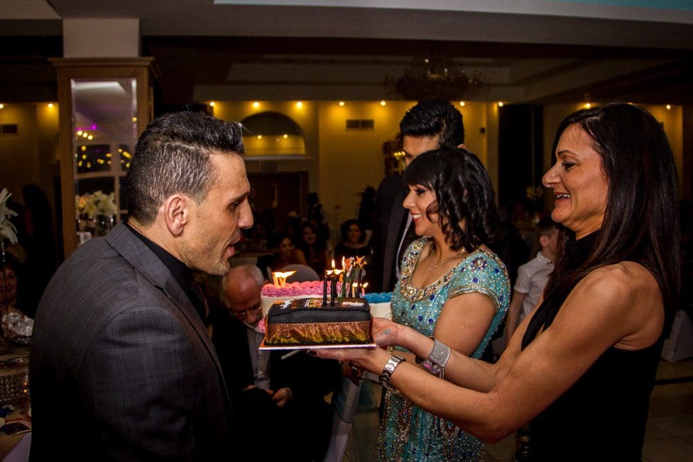A man leans towards a birthday cake with lit candles, held by a smiling woman. Other people stand nearby, smiling and celebrating indoors—capturing the perfect moment for a Birmingham event photographer.