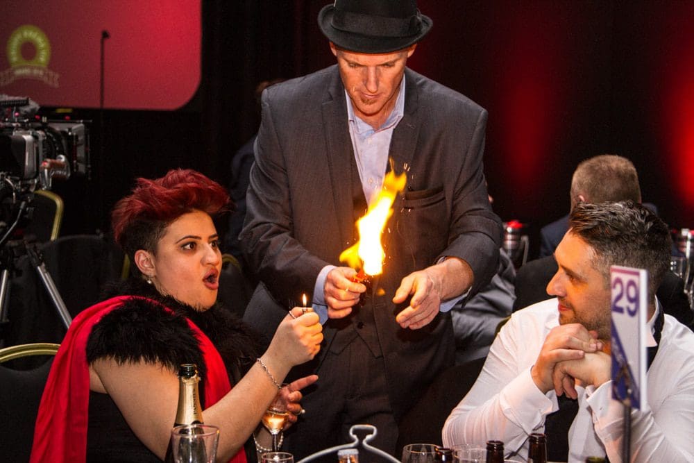 A magician in a suit performs a fire trick at a dining table, surprising a woman with red hair and a man who are watching closely, as captured by a skilled Birmingham event photographer.