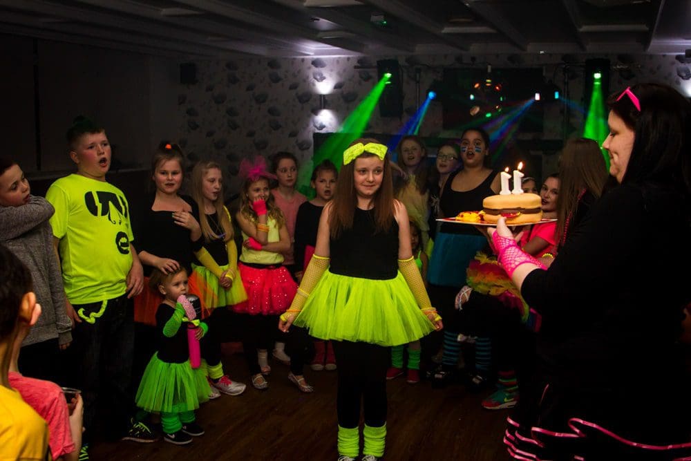 A girl in a neon yellow tutu stands as a woman brings her a birthday cake with candles. Children and adults in colourful neon outfits gather round, smiling and watching. The room is dimly lit with party lights, capturing the perfect moment for any Birmingham event photographer.