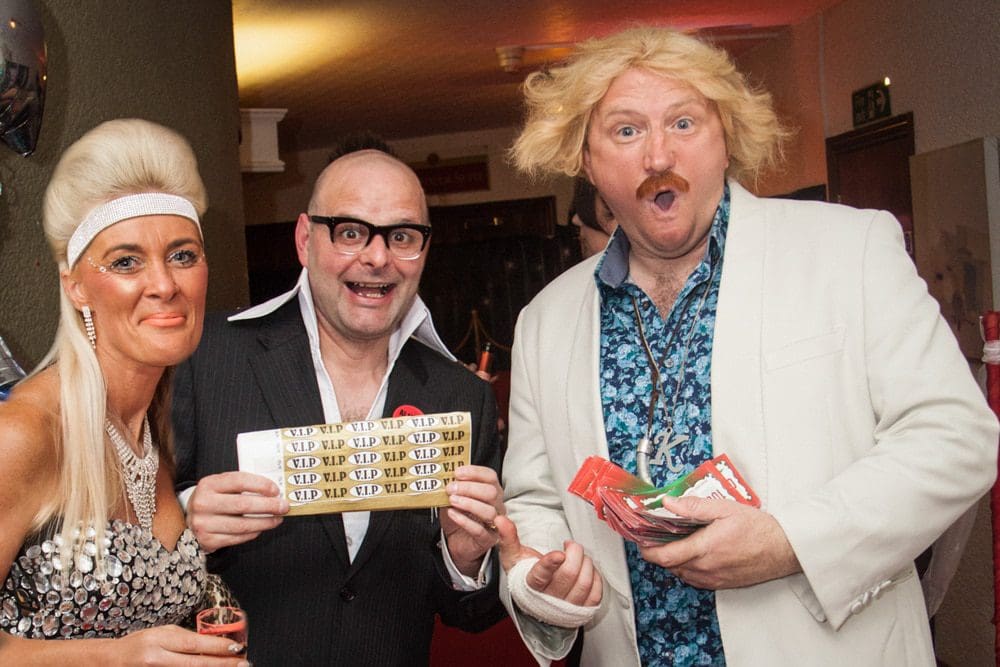 Three people in flashy costumes pose indoors; one woman in a sparkling dress, and two men, one bald with glasses holding VIP tickets, the other with messy blond hair and a moustache holding snacks, all smiling excitedly. Captured by a skilled Birmingham event photographer, this lively group radiates enthusiasm and fun.