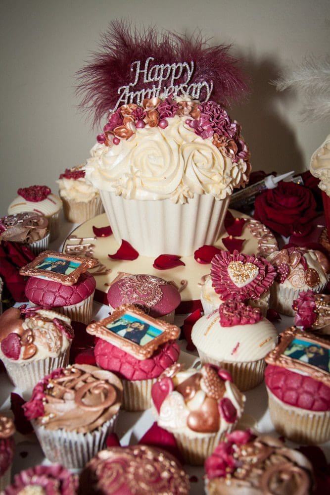 A large cupcake with Happy Anniversary topper is surrounded by smaller cupcakes decorated with red, gold, and white details, including edible photos, pearls, and roses—perfect for capturing by a Birmingham event photographer.