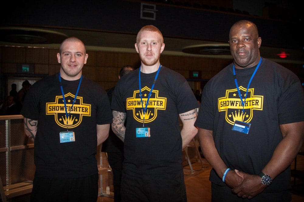 Three security staff in matching black SHOWFIGHTER shirts and blue lanyards stand side by side indoors, facing the camera and smiling—captured perfectly by a Birmingham adult birthday photographer.