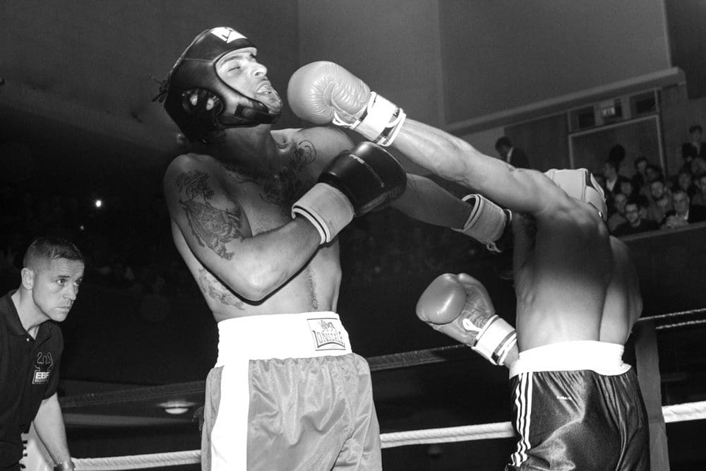 A black-and-white photo, captured by a skilled Birmingham event photographer, shows a boxer landing a punch to his opponent’s face in a ring, whilst a referee watches closely. The audience is visible in the background.