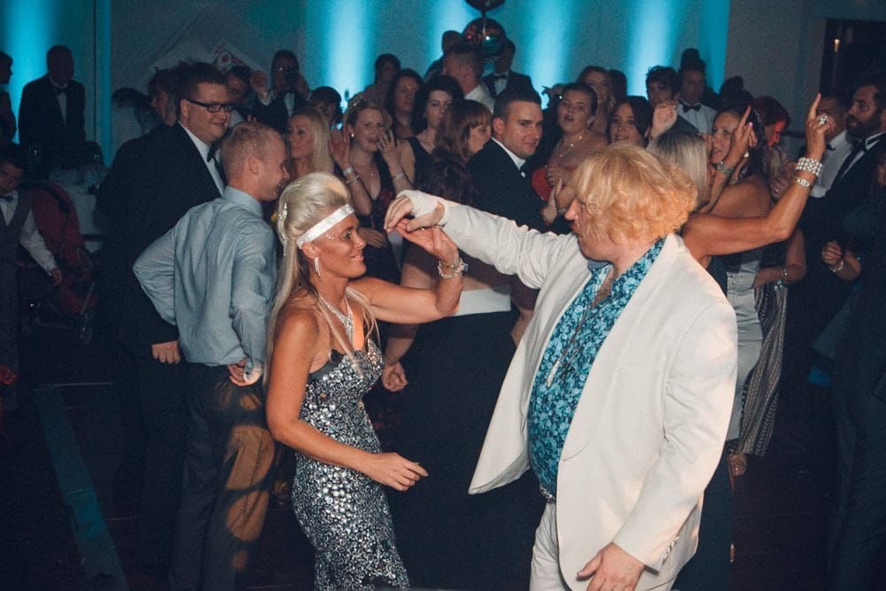 A man and woman in flashy outfits dance together at a lively party, surrounded by a crowd of smiling people in formal attire. Blue lights illuminate the background, perfectly captured by a skilled Birmingham event photographer.