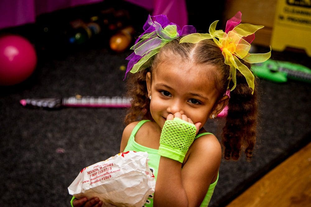 A young girl with colourful bows in her hair smiles shyly, wearing a neon green outfit and fingerless glove, holding a crumpled paper bag at what appears to be a party—captured through the lens of a skilled Birmingham event photographer.