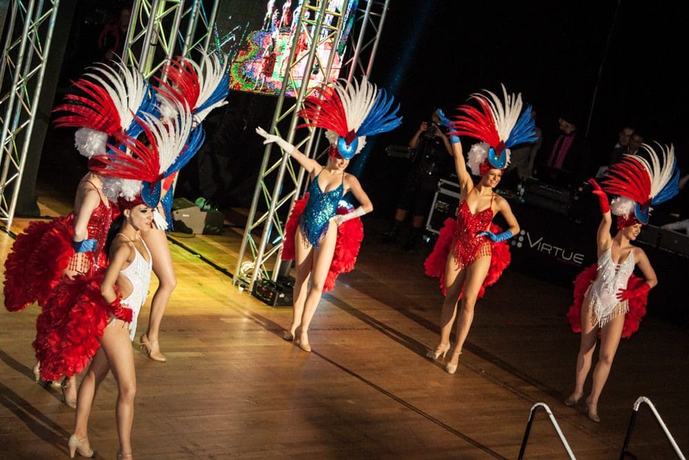Six dancers in red, white, and blue costumes with large feathered headpieces perform on a stage with spotlights and stage structures in the background, captured vividly by a Birmingham event photographer.
