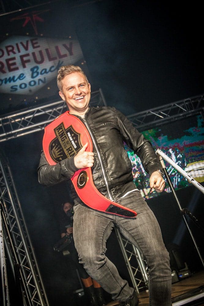 A smiling man in a black jacket and jeans holds a red wrestling-style championship belt while walking down steps on a stage, expertly captured by a Birmingham event photographer, with lights and a Drive Carefully sign in the background.