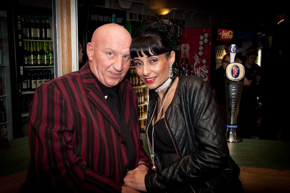 A man in a red and black striped suit stands next to a woman in a black jacket at a bar, both smiling at the camera. Captured by a Birmingham event photographer, bottles and a beer pump are visible in the background.