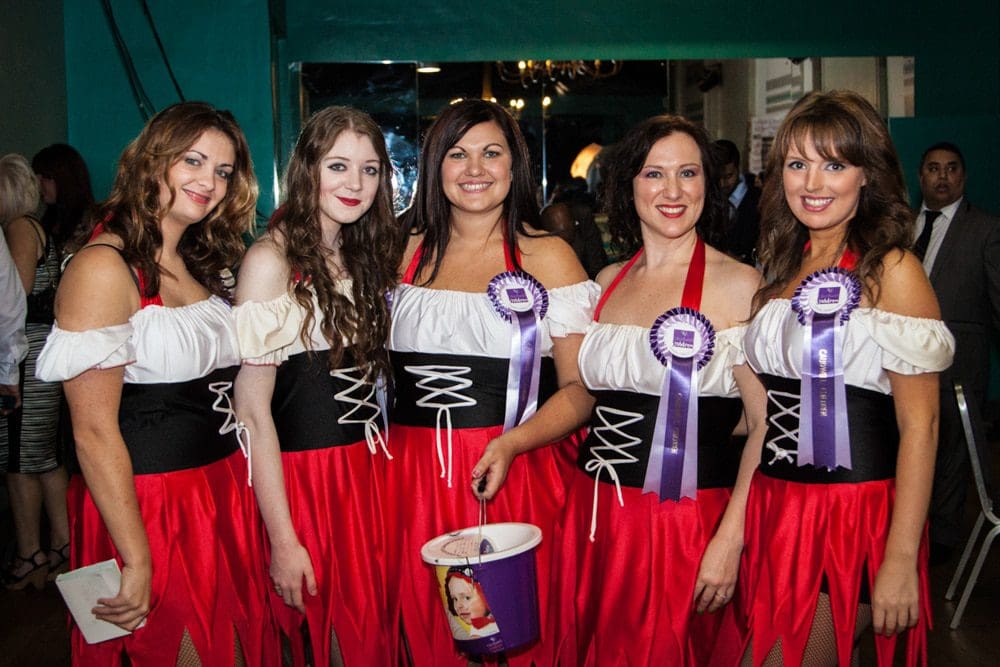 Five women wearing matching red, white, and black costumes stand together. Three wear purple rosettes and hold a collection bucket. Captured by a Birmingham event photographer, they are smiling, with a few people visible in the background.