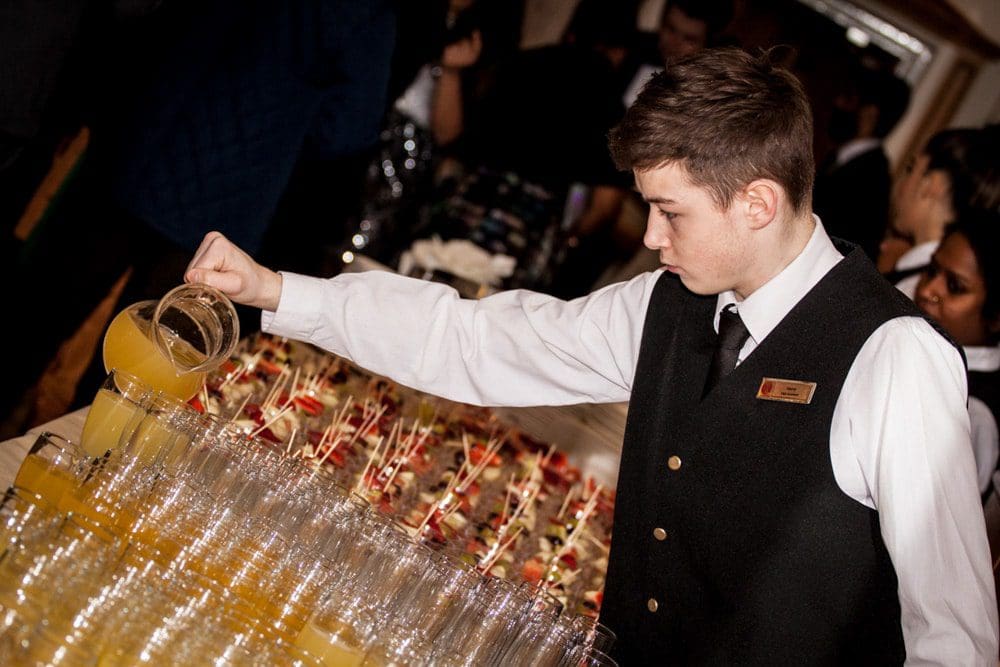 A waiter in a black waistcoat pours orange juice into rows of clear glasses at a crowded event, with other servers and guests in the background—captured with the keen eye of a Birmingham event photographer.